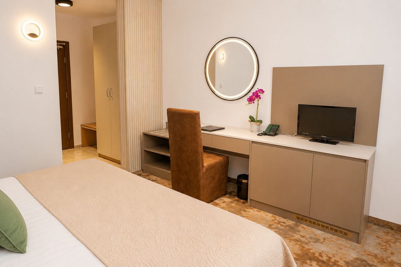 A hotel room with a bed, a desk with a chair, a television, a mirror, and a small potted plant.