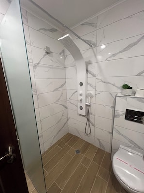 Une salle de bain moderne avec une douche à parois de verre, des carreaux de marbre, des toilettes avec un abattant et une petite plante posée sur l’étagère.