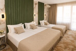 A hotel room with two beds, a green headboard, a small round table with a vase of flowers, and a large window with curtains.