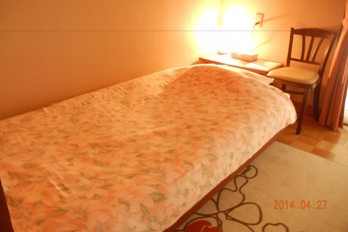 Une petite chambre avec un lit simple, une couvre-lit à motifs floraux, une chaise et une table de chevet avec une lampe.