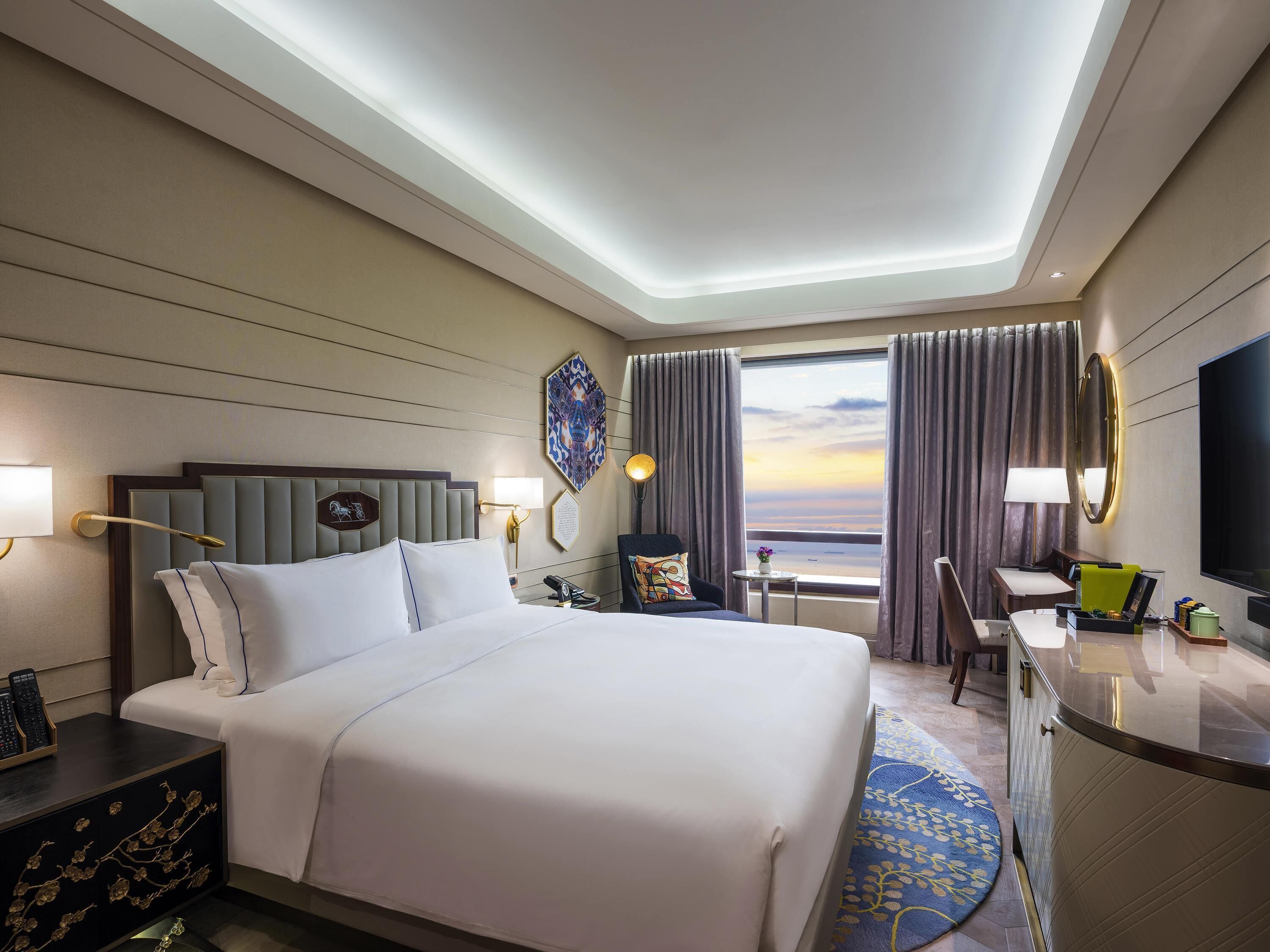 Une chambre d’hôtel comprenant un lit, une table de chevet, un bureau, une chaise et une grande fenêtre avec des rideaux.
