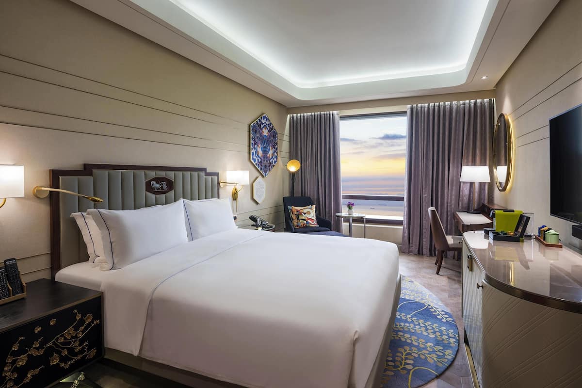 A hotel room with a bed, bedside table, desk, chair, and a large window with curtains.