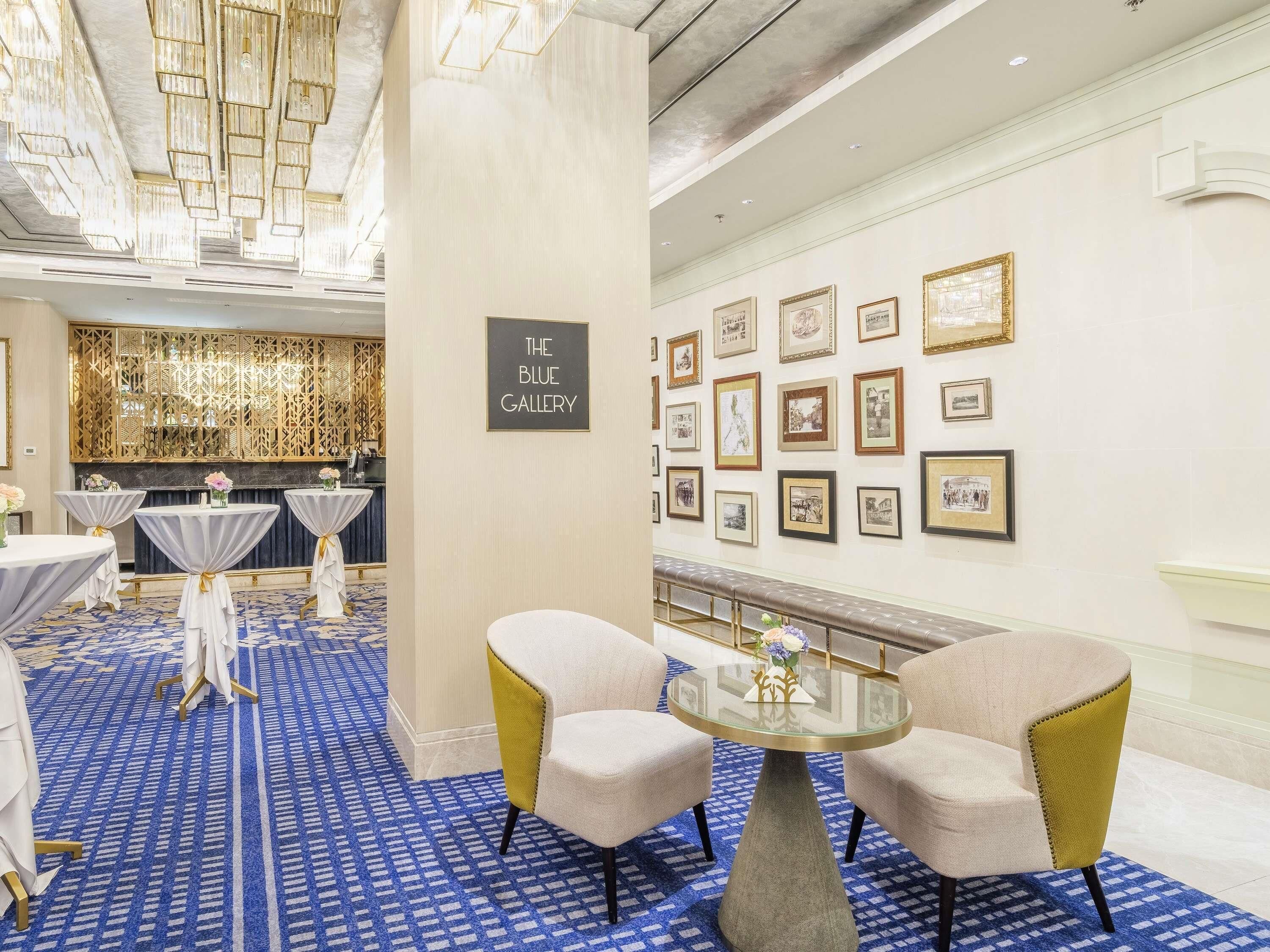 A hotel lobby with a blue patterned carpet, round tables with white tablecloths, a glass coffee table, and a wall displaying framed pictures.