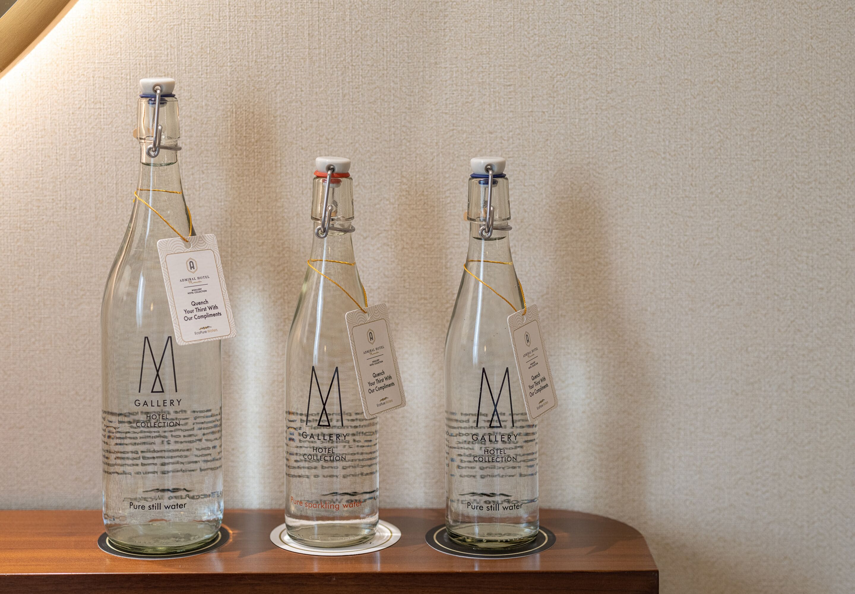 Tres botellas de vidrio con la etiqueta 'Gallery Hotel Collection' y etiquetas, sobre una superficie de madera frente a una pared texturizada.