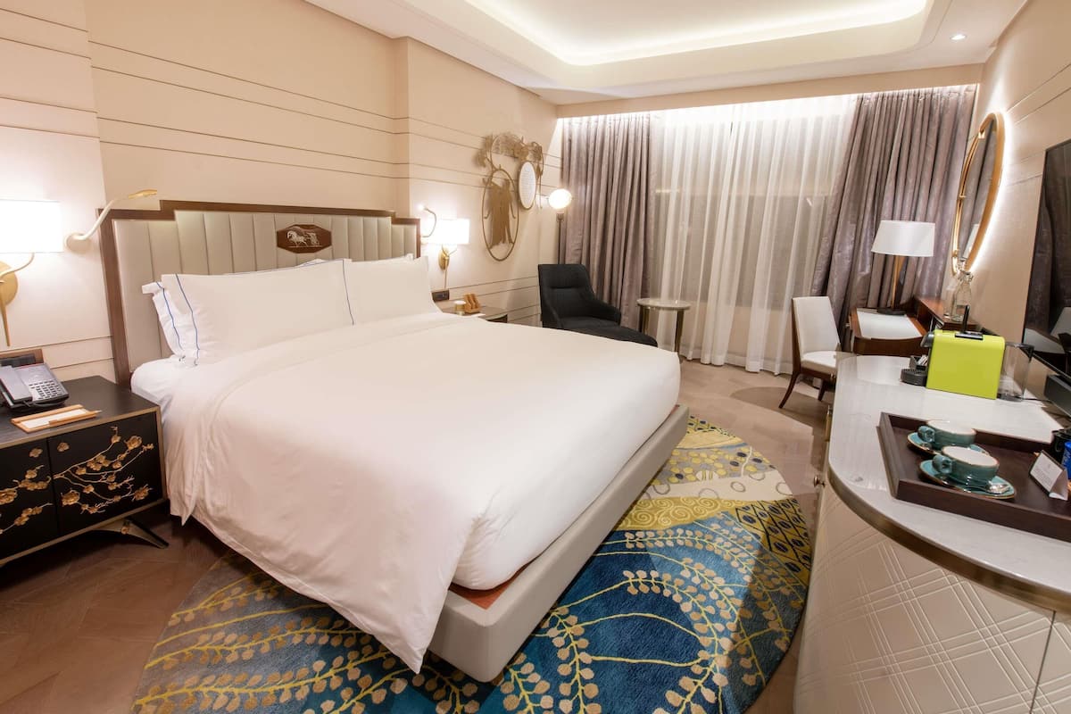 A hotel room with a large bed, bedside tables, a desk, a chair, a mirror, and a window with curtains.