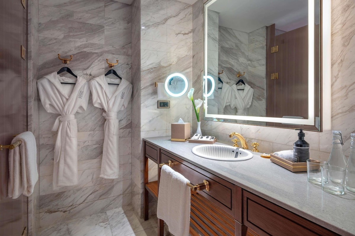 A bathroom with marble walls, a large mirror, two white robes hanging, a vanity with a sink, and a towel rack.