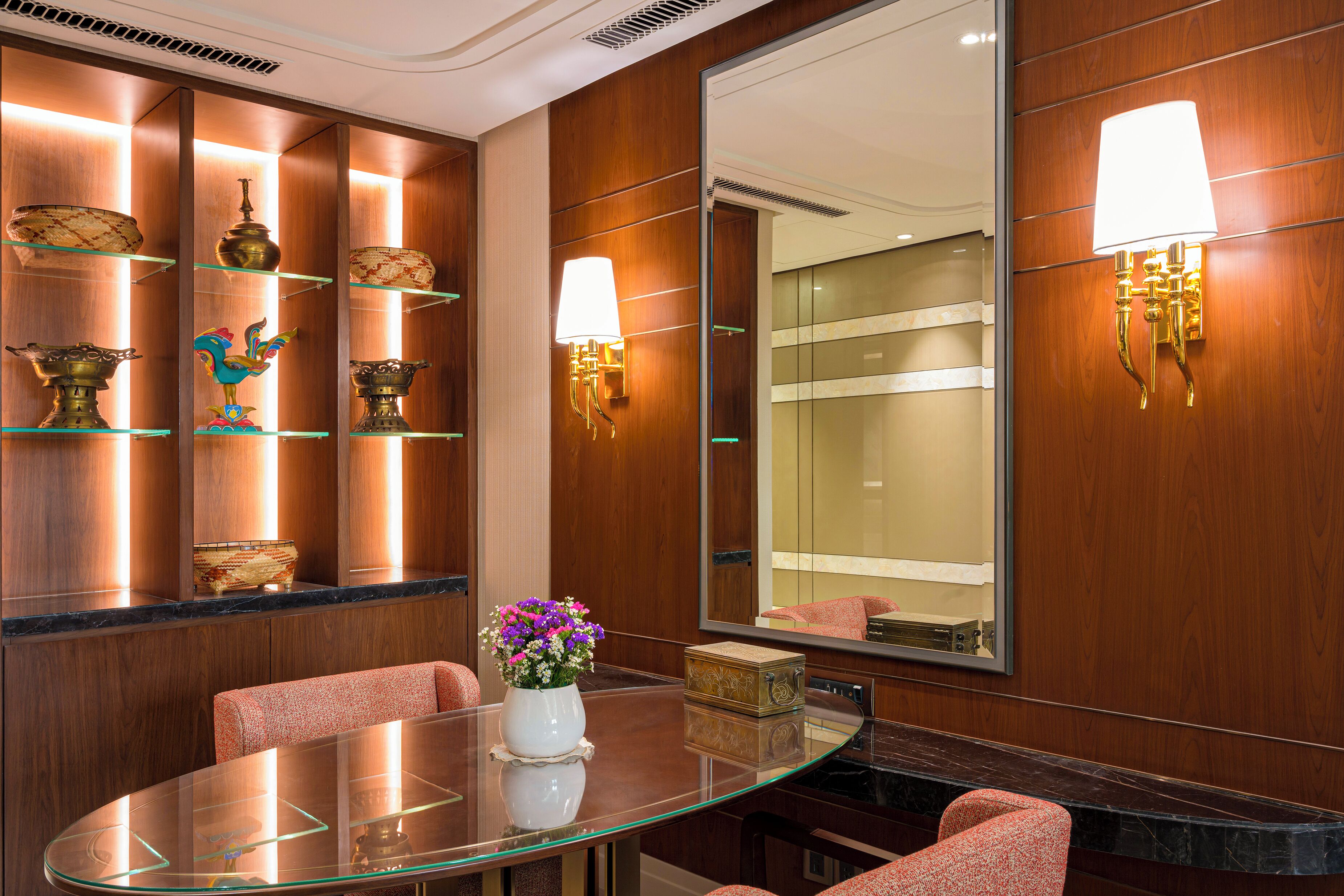A hotel lobby with a glass table, red upholstered chairs, decorative items on shelves, and a large mirror.