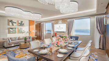 A hotel dining room with a large table set for six, surrounded by comfortable seating, a chandelier, and a large window with a view.