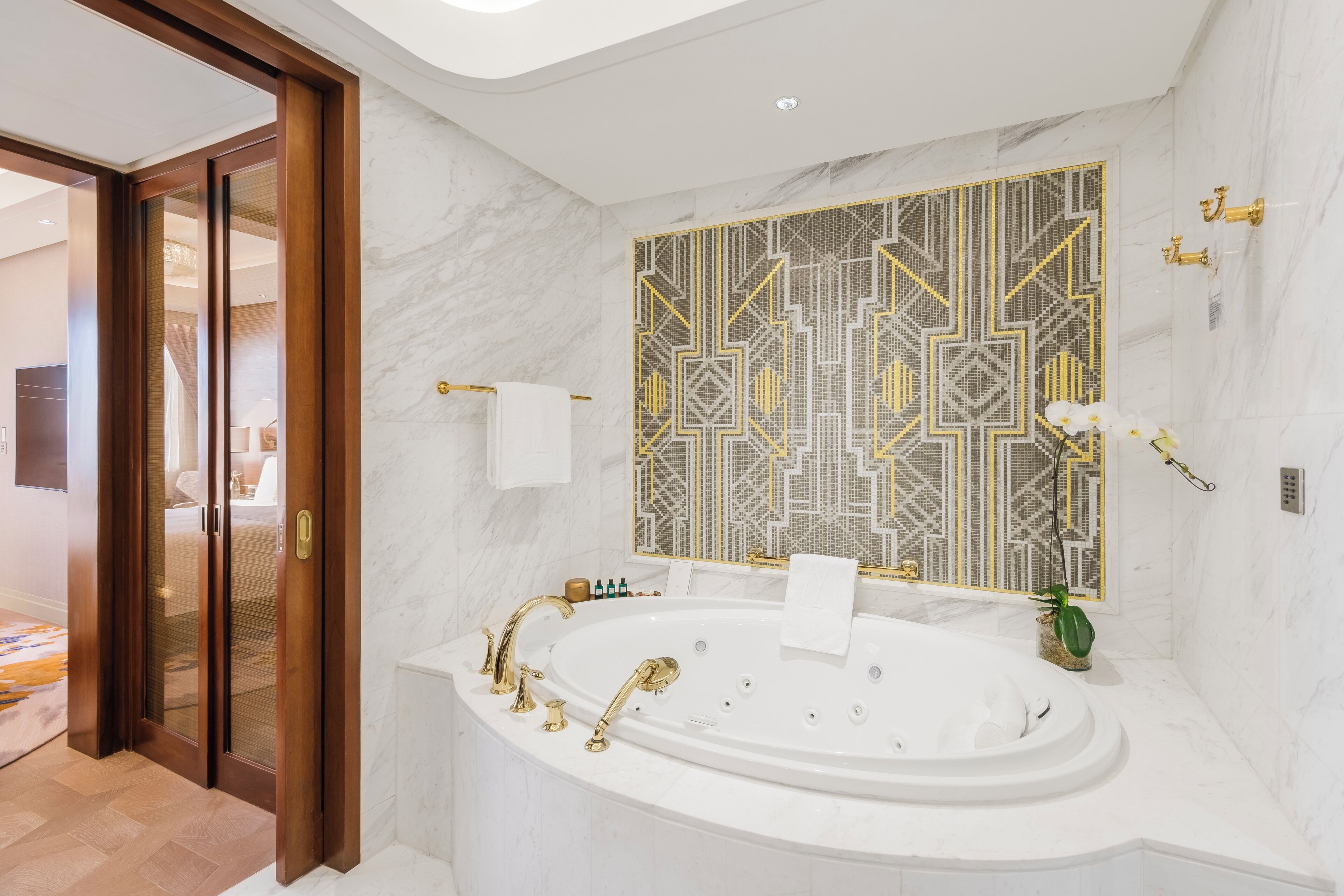 Un baño moderno con una bañera independiente, un espejo grande con un reverso de azulejos geométricos y una puerta de madera que da a otra habitación.