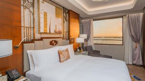 Habitación de hotel con una cama grande, un escritorio con teléfono, una silla, una lámpara de escritorio, una ventana con cortinas y obras de arte en la pared.