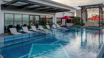 Une piscine à débordement située sur le toit, avec des chaises longues et des parasols, un bar entièrement vitré et une vue imprenable sur les toits de la ville.