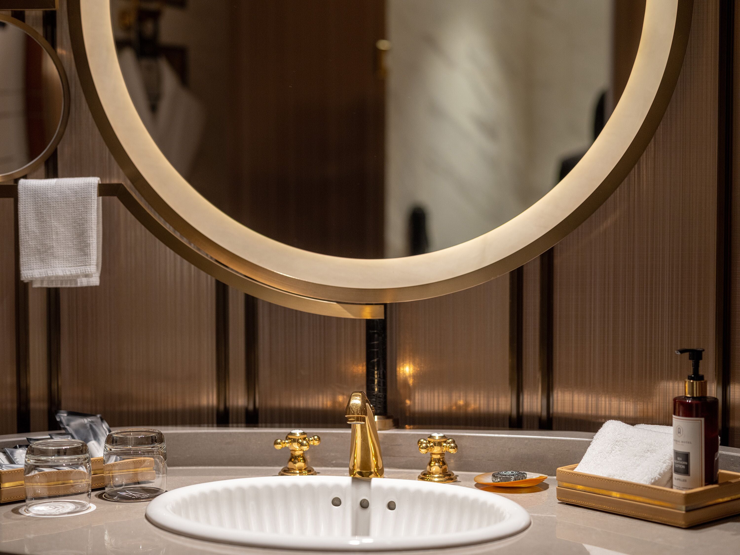 Une salle de bain avec un lavabo blanc, des robinets dorés et un grand miroir. On y trouve des articles de toilette sur le comptoir et un porte-serviettes au-dessus du miroir.