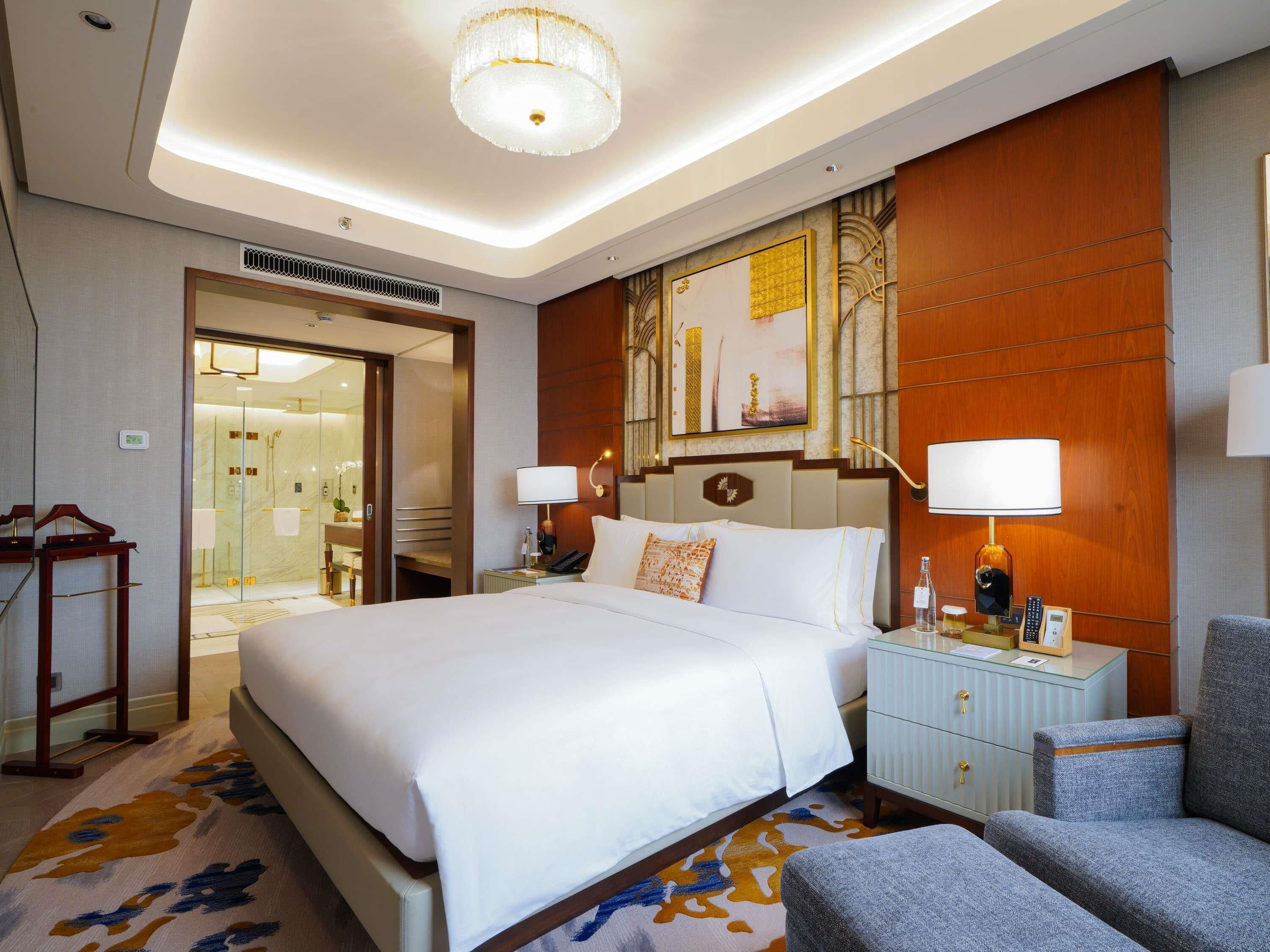 A hotel room with a large bed, a bedside table with a lamp, a chair, a wardrobe, and a bathroom visible through an open door.