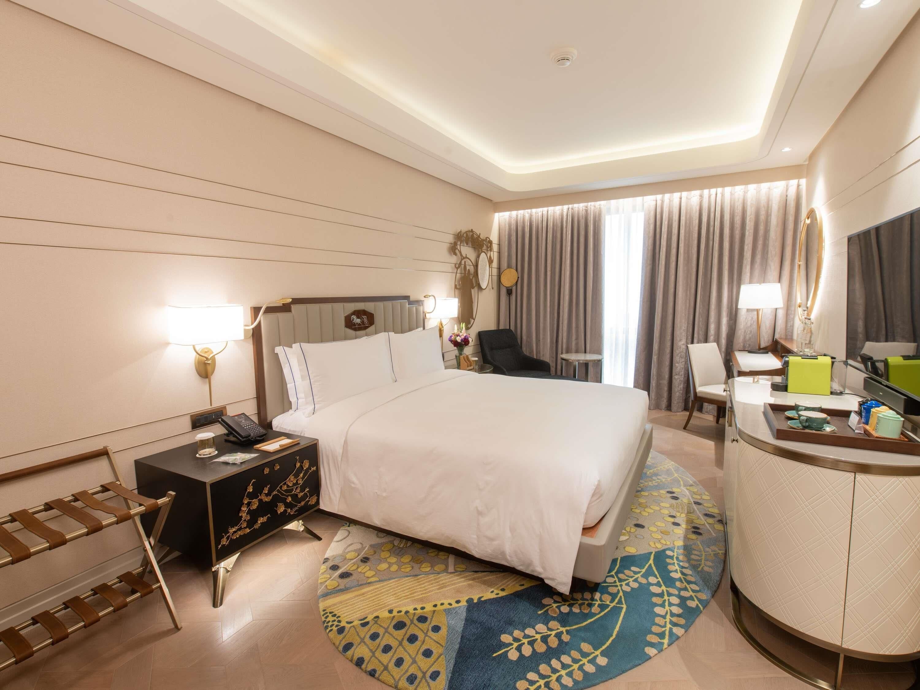 A hotel room with a large bed, bedside tables, a desk, a chair, a round coffee table, and a mirror.