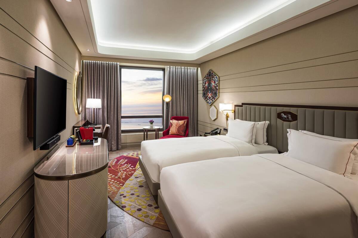 A hotel room with a flat-screen TV, two beds, a desk, a chair, a small table, and a view of the sea.