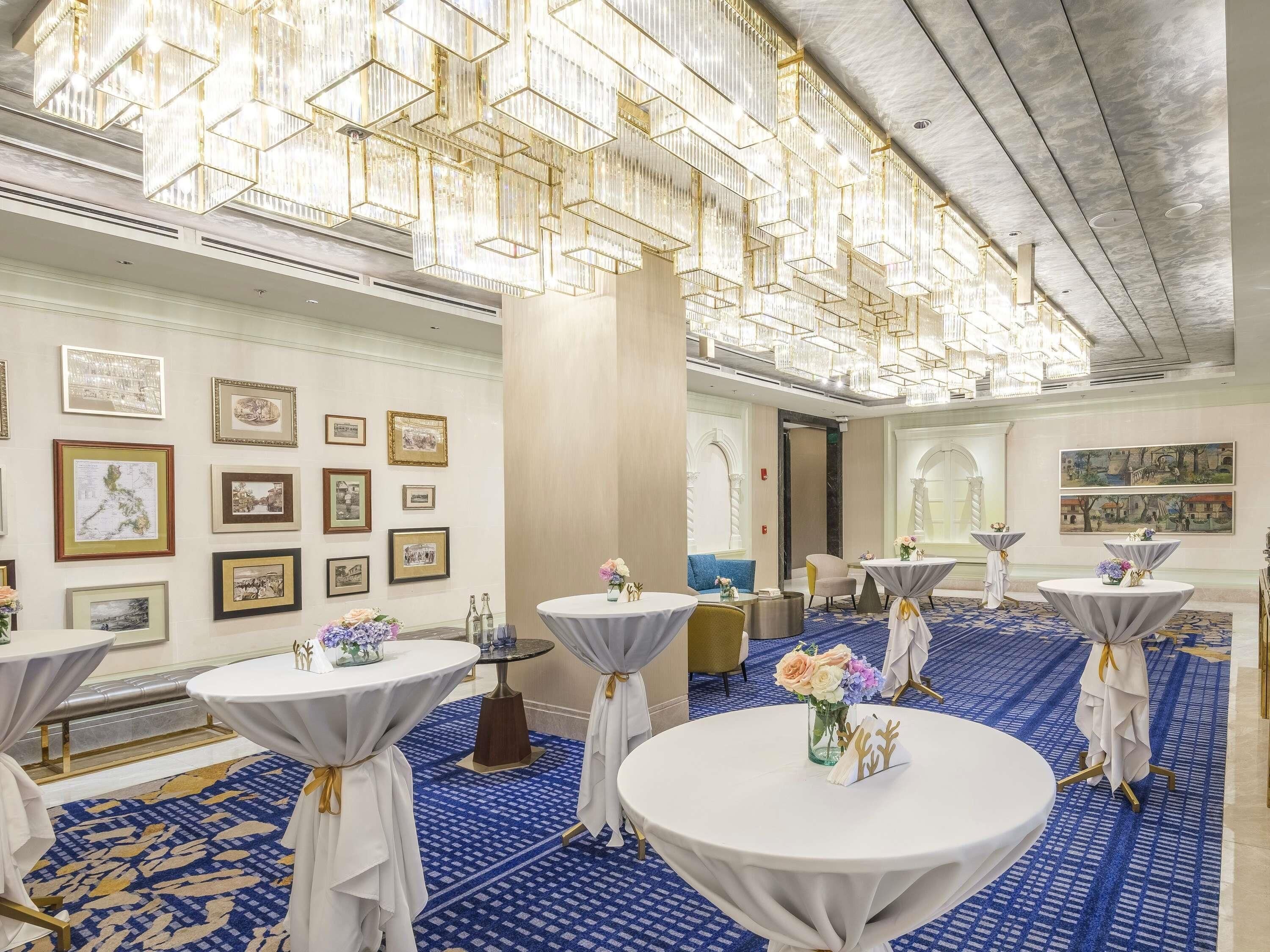 A hotel room with round tables, a chandelier, framed pictures on the wall, and a blue patterned carpet.
