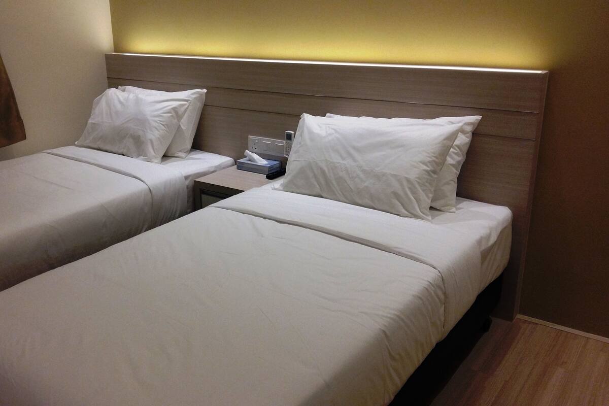 A hotel room with two single beds, each with white linens and pillows, a bedside table with a remote control, and a wall-mounted light.