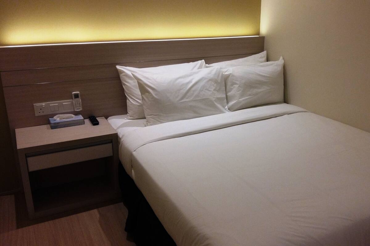 A single bed with white linens, a bedside table with a tissue box, a remote control, and a phone charger.