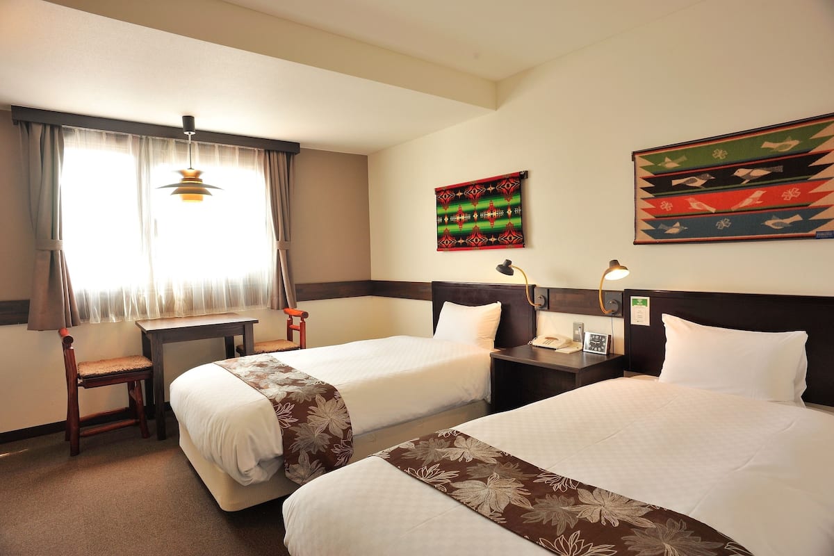Ein Hotelzimmer mit zwei Betten, einem Schreibtisch, einem Stuhl und einem Fenster mit Vorhängen.