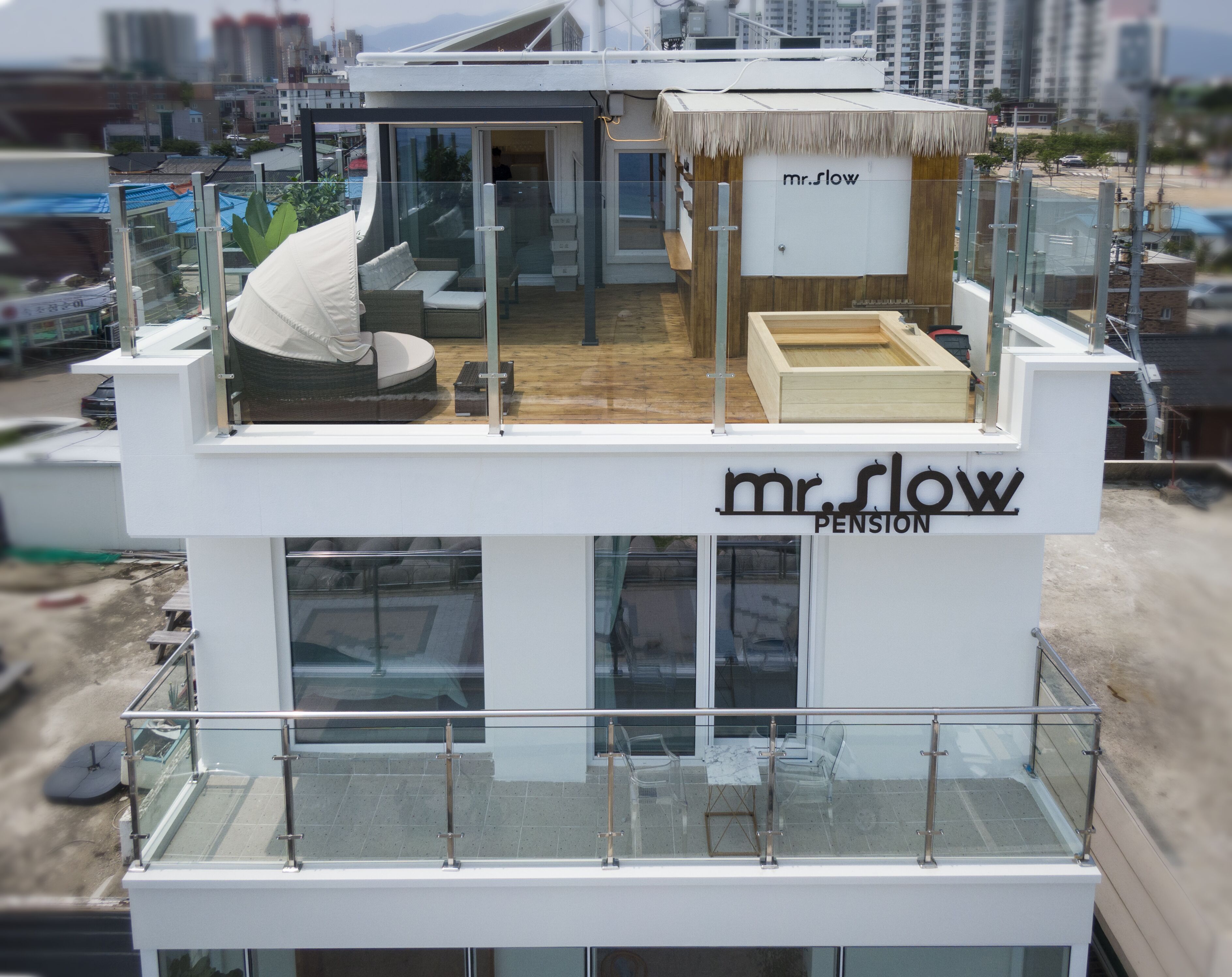 A modern building with a glass balcony, a lounge area with a thatched roof, and the name "Mr. Slow Pension" on the facade.