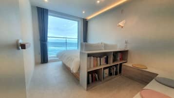 A modern bedroom with a large window overlooking the ocean, a bed, a bedside bookshelf, and a small table with a chair.