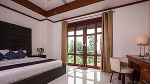 A hotel room with a large bed, a desk, a chair, and a view of greenery through the window. - NB Villa Sienna (Koh Samui)