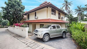 A two-story house with a red-tiled roof, a silver pickup truck parked in front, and lush greenery surrounding the property. - NB Villa Sienna (Koh Samui)