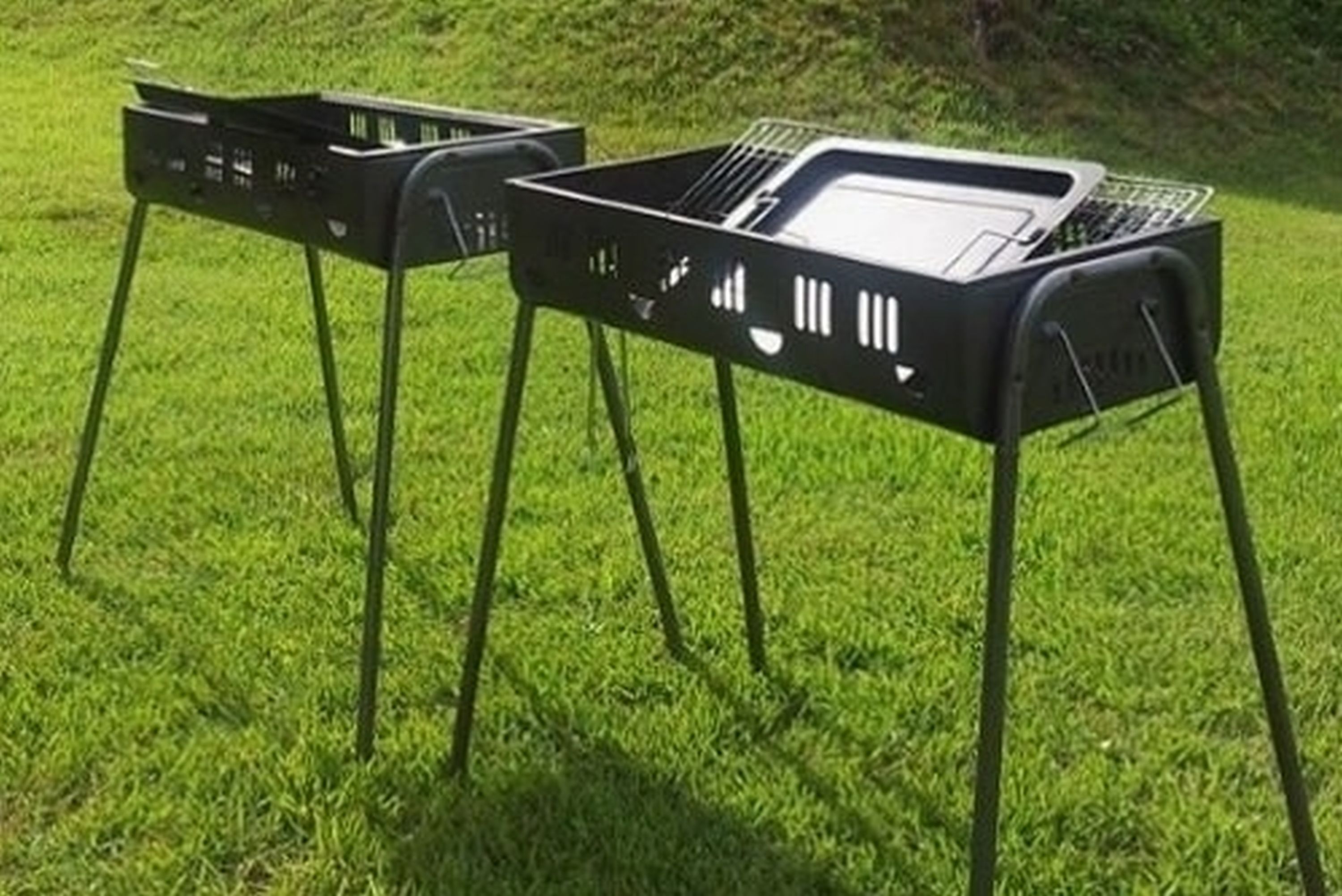 Two portable black barbecue grills on a grassy lawn.