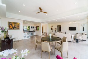 A spacious living area with a round glass table, beige chairs, a ceiling fan, and a TV mounted on the wall.