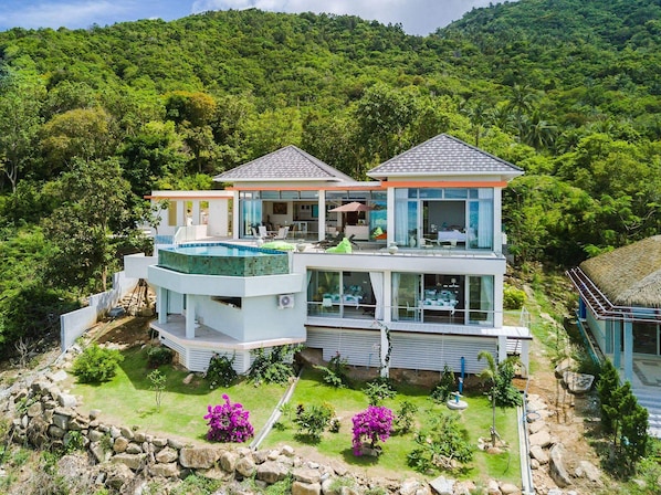 A modern two-story house with a swimming pool, surrounded by lush greenery and hills.