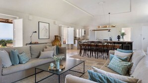 En rummelig opholdsstue med sofa, sofabord og spisebord. I baggrunden er der et køkken med skabe og en hængende lampe.