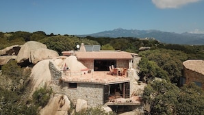 Ein Steinhaus mit Ziegeldach, einer Terrasse mit Gartenmöbeln und Blick auf Berge im Hintergrund.
