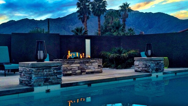 A pool area with stone fire pits and lounge chairs, surrounded by palm trees and mountains. - Fantastic Views and Reviews! Bright, Relaxing Oasis! (Palm Springs)