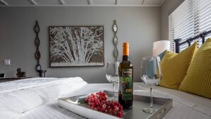 A neatly made bed with white linens, a tray with grapes and a bottle of wine, and a painting of trees on the wall.