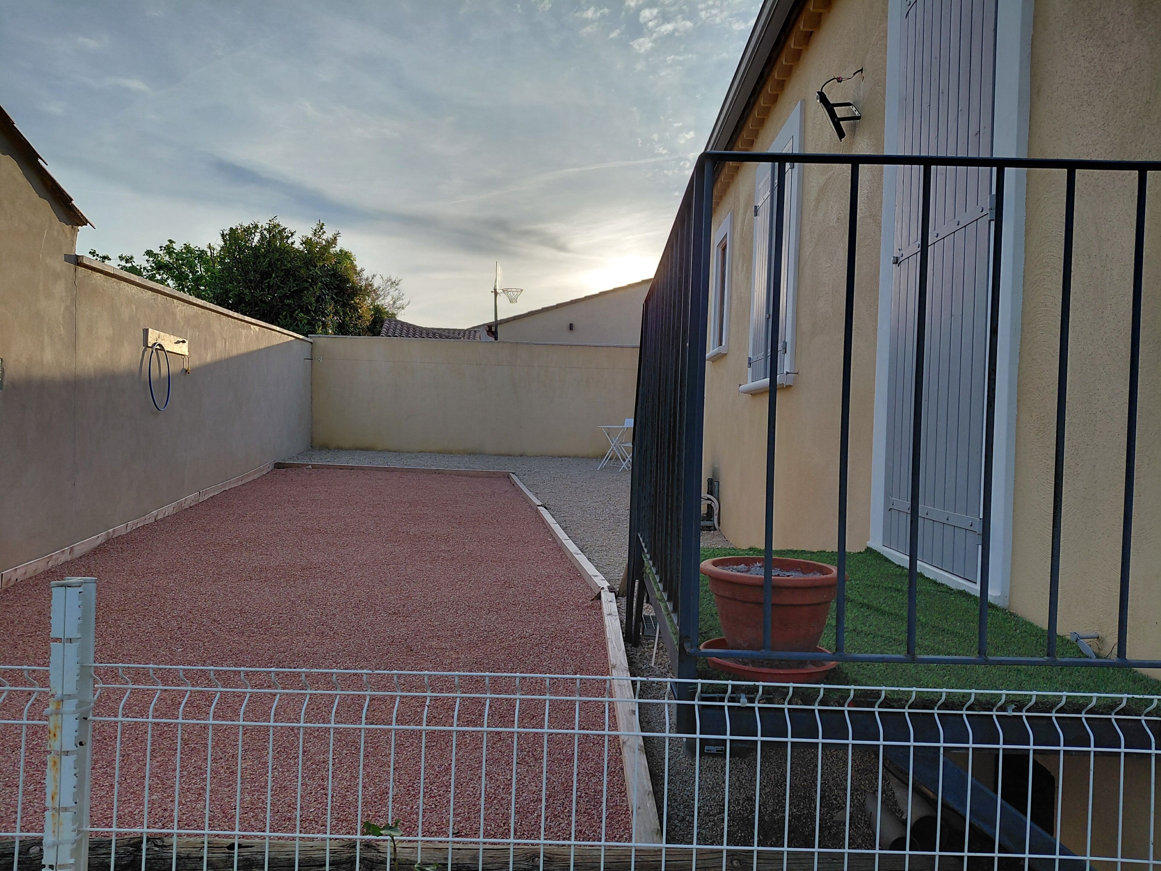 Eine Dachterrasse mit rotem Kiesbelag, einem Metallzaun, einem Topfpflanzen und einem Gebäude mit einer beigen Wand und Fenstern.