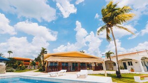A pool area with lounge chairs, a thatched-roof building, and palm trees. - Costa A6 Beautiful Beachfront Property (Cozumel)