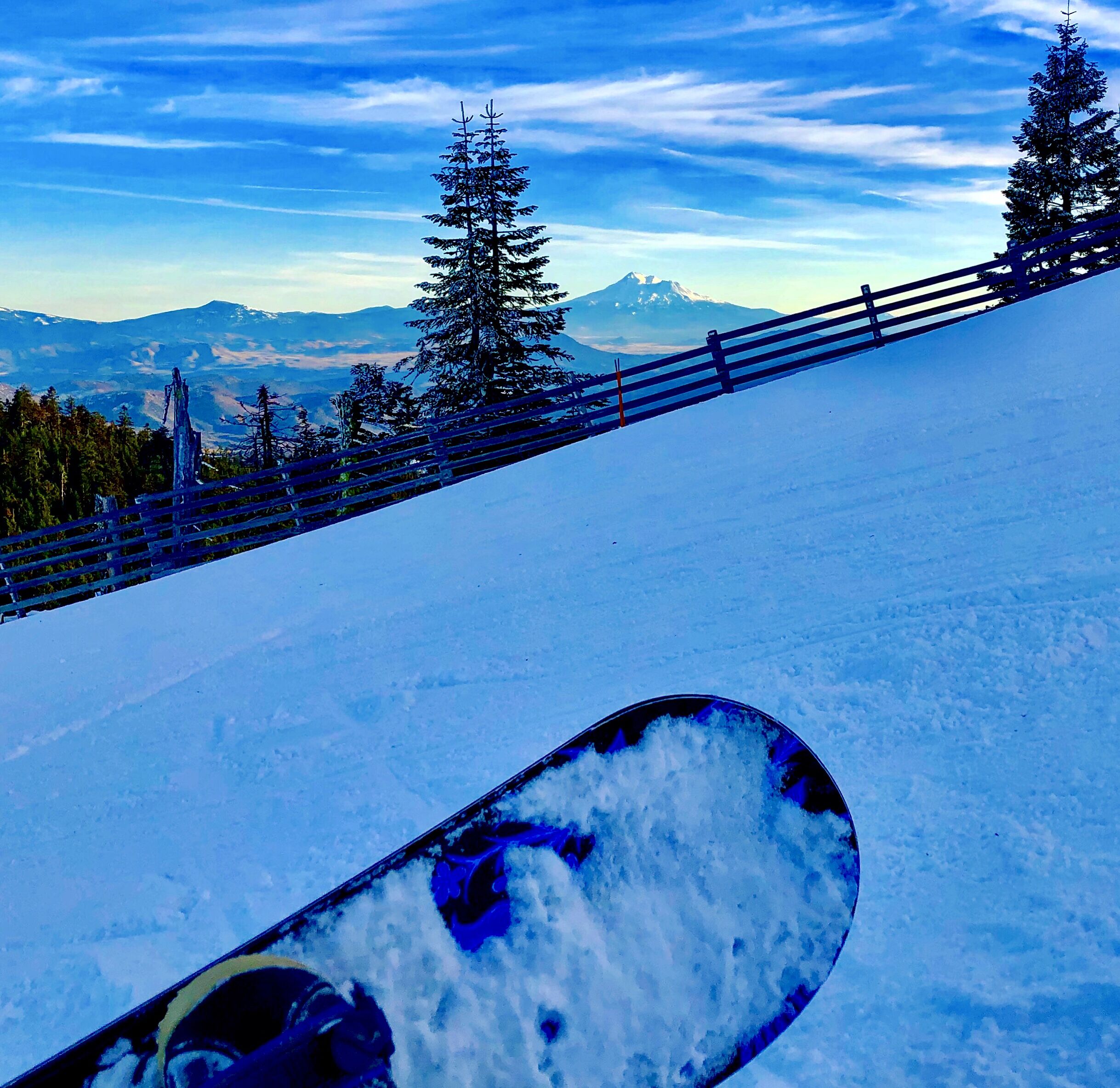 A snowboard on a snowy slope with a wooden fence and trees in the background.