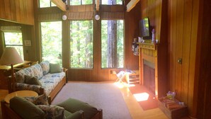 A cozy cabin interior with a sofa, chairs, a small table, a fireplace, and large windows offering a view of the forest. - Dog-Friendly, Secluded Redwoods Getaway With Private Hot Tub (Sea Ranch)