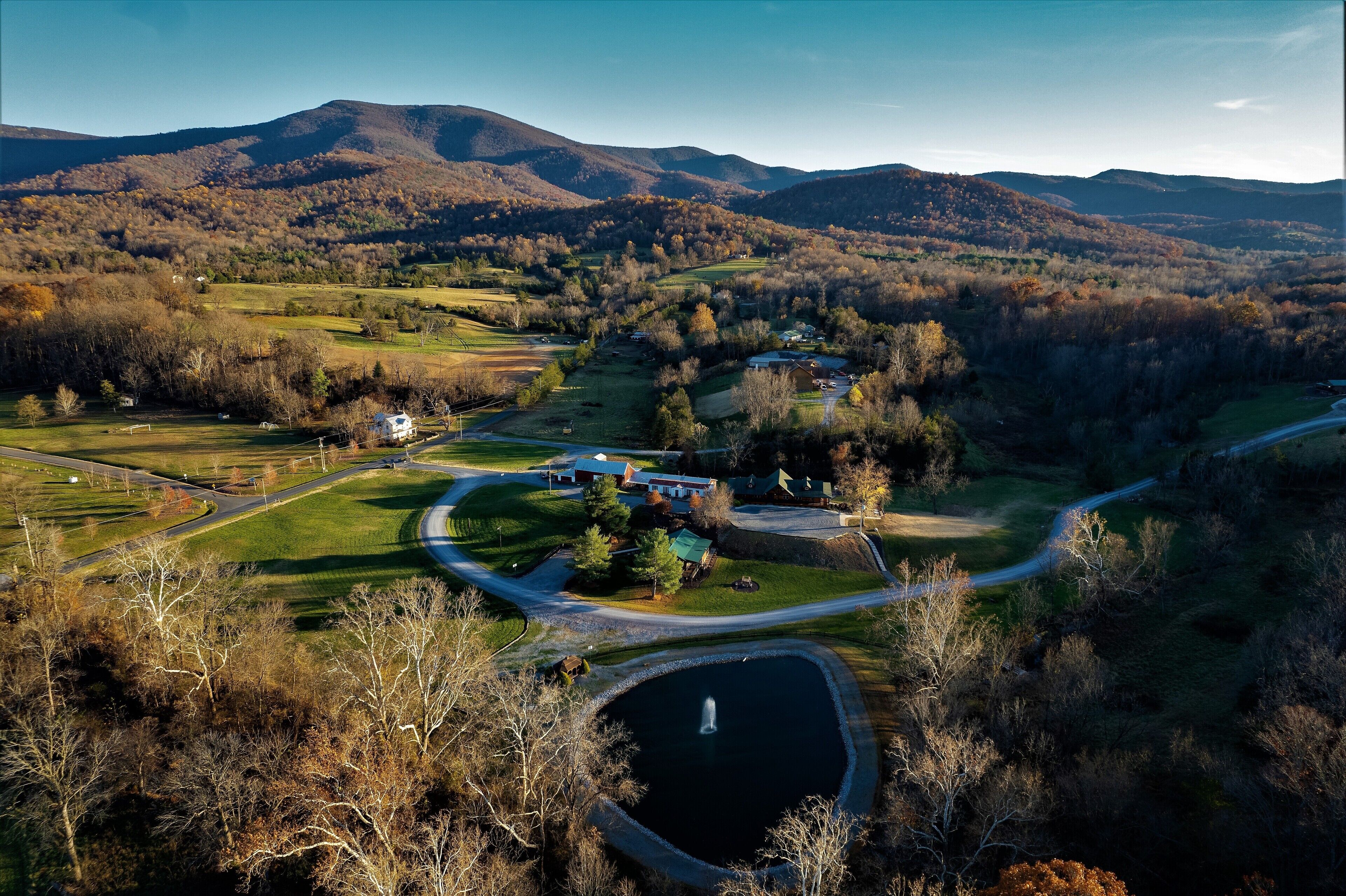 Un paysage rural avec une route sinueuse, un étang et un petit bâtiment entouré d’arbres et de collines.