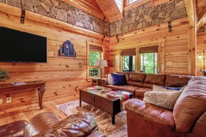A log cabin interior with a stone ceiling, wooden walls, a leather sofa, a coffee table, and a television.
