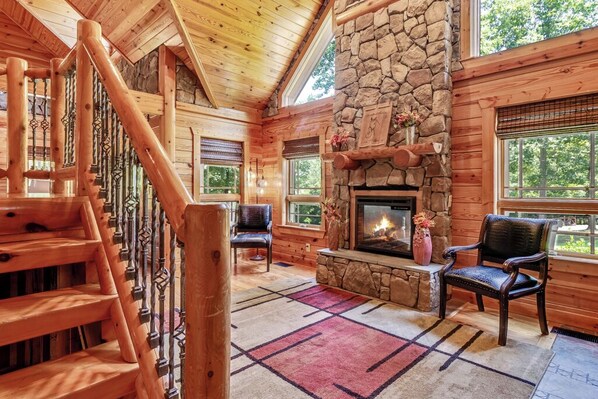 A log cabin interior with a stone fireplace, wooden staircase, and large windows.