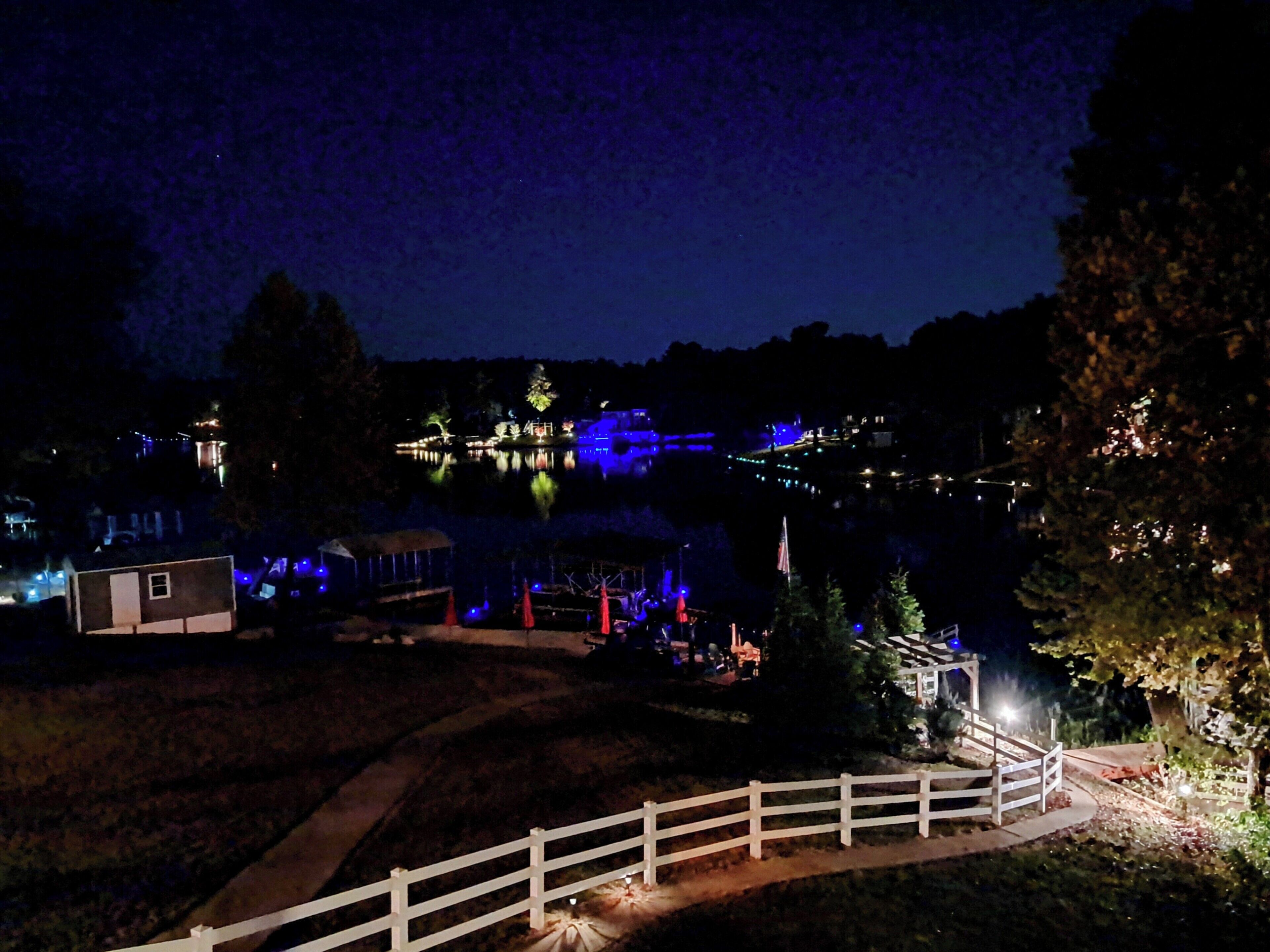A nighttime view of a lakeside area with illuminated buildings and a white fence.