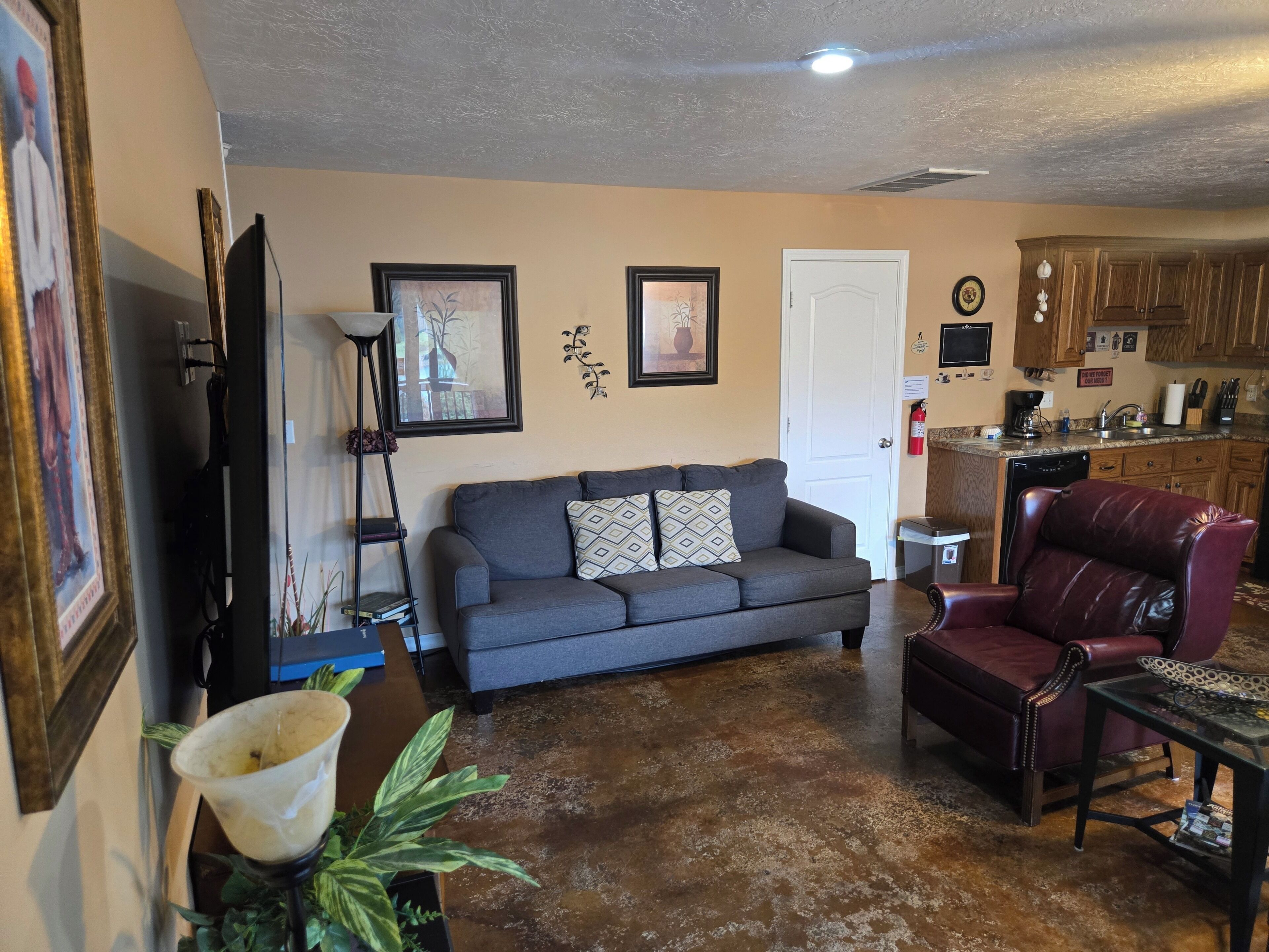 A living room with a gray sofa, a red leather chair, a coffee table, and a kitchen area with wooden cabinets and a dishwasher.