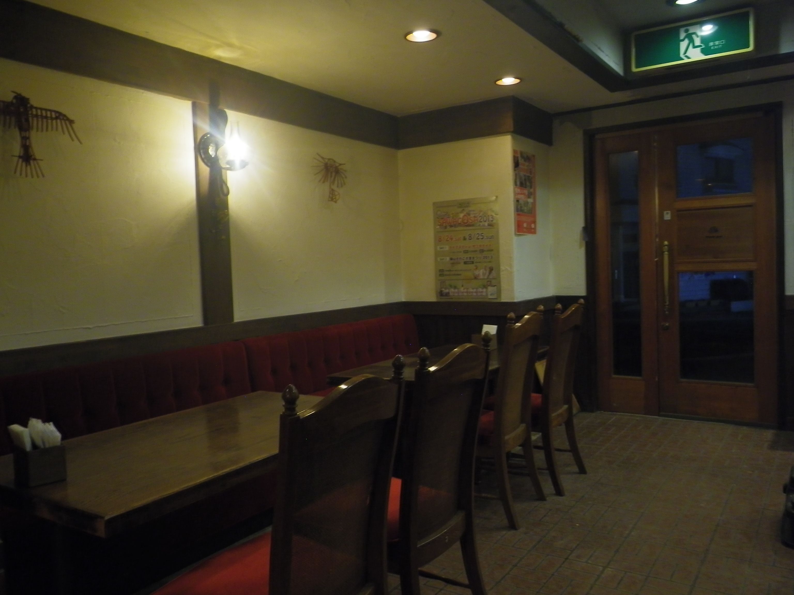 En restaurantindretning i dæmpet belysning med træborde og siddepladser, en vægmonteret lampe og en dør med et glaspanel.