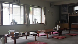 Um quarto tradicional japonês com tatames, uma mesa baixa com chaleira, xícaras e tigelas, e vista para o exterior através de grandes janelas.