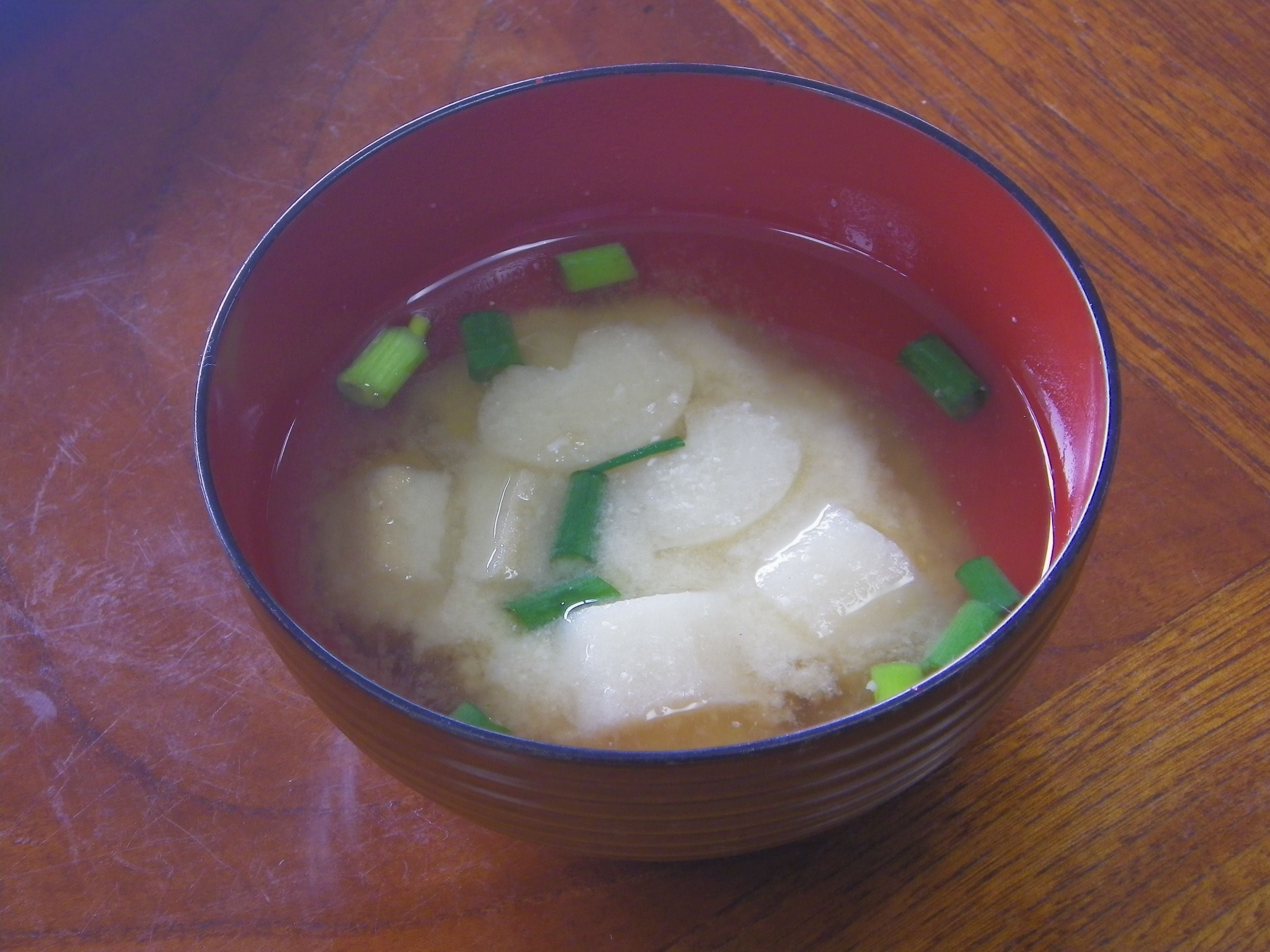 A bowl of clear soup with green onions and white cubes, possibly tofu, on a wooden table.