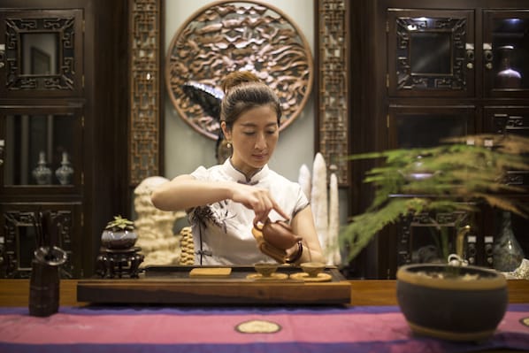 Una persona realizando una ceremonia del té en una sala tradicional con muebles de madera y arte decorativo en las paredes.