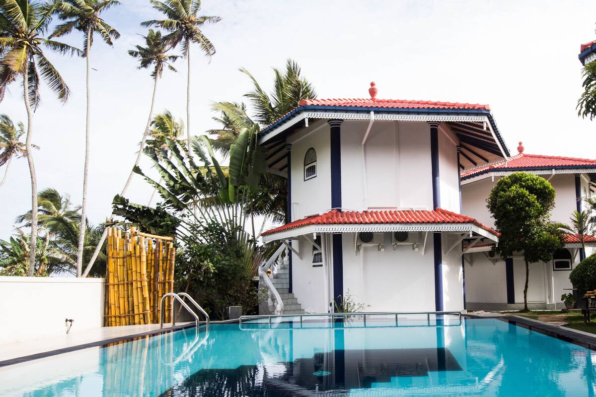 A swimming pool with a red-tiled roof, surrounded by palm trees and a white building with blue accents.