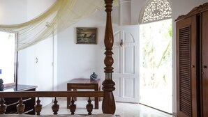 A bedroom with a four-poster bed, a wooden bench, a small table, and a door leading to a bright room with a view of greenery. - Neptune Story Hotel (Weligama)