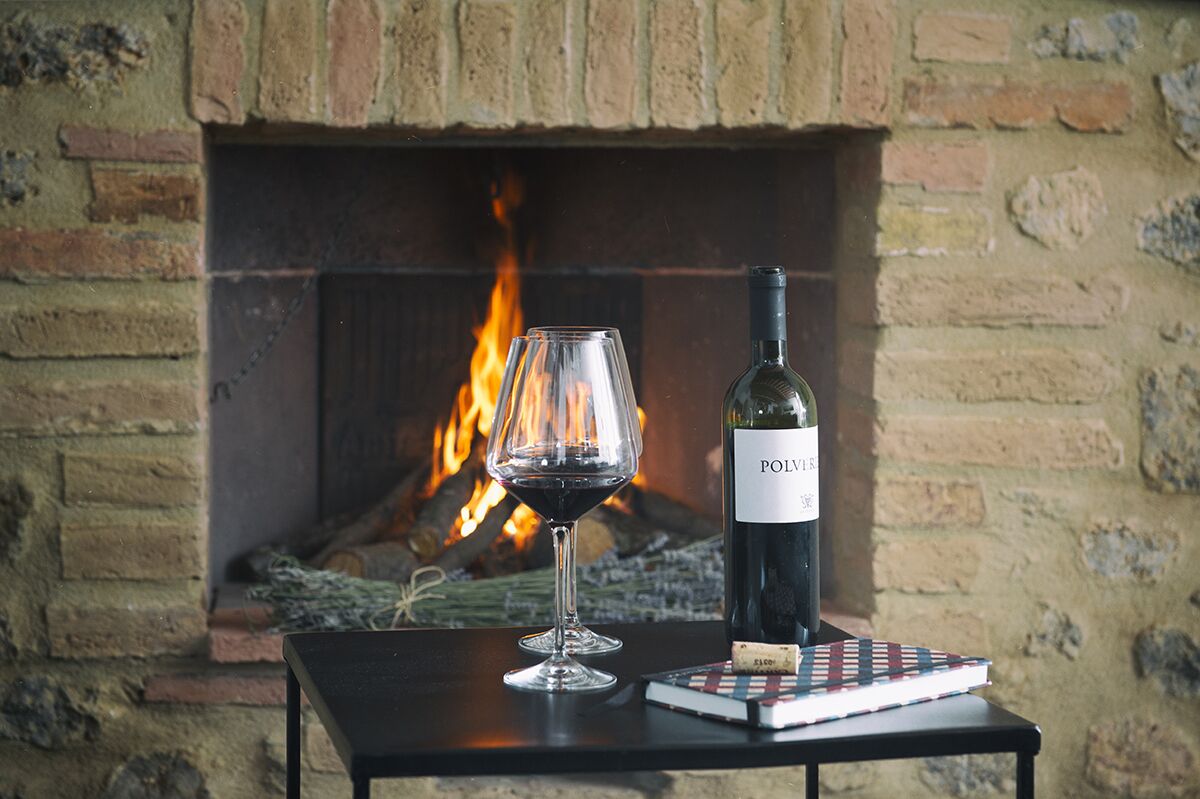 Una mesa con una botella de vino, una copa y un libro frente a una chimenea.